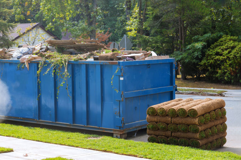 Lawn grass in rolls on pallets against of the street the rolled grass lawn is ready for laying and loaded dumpster near a construction